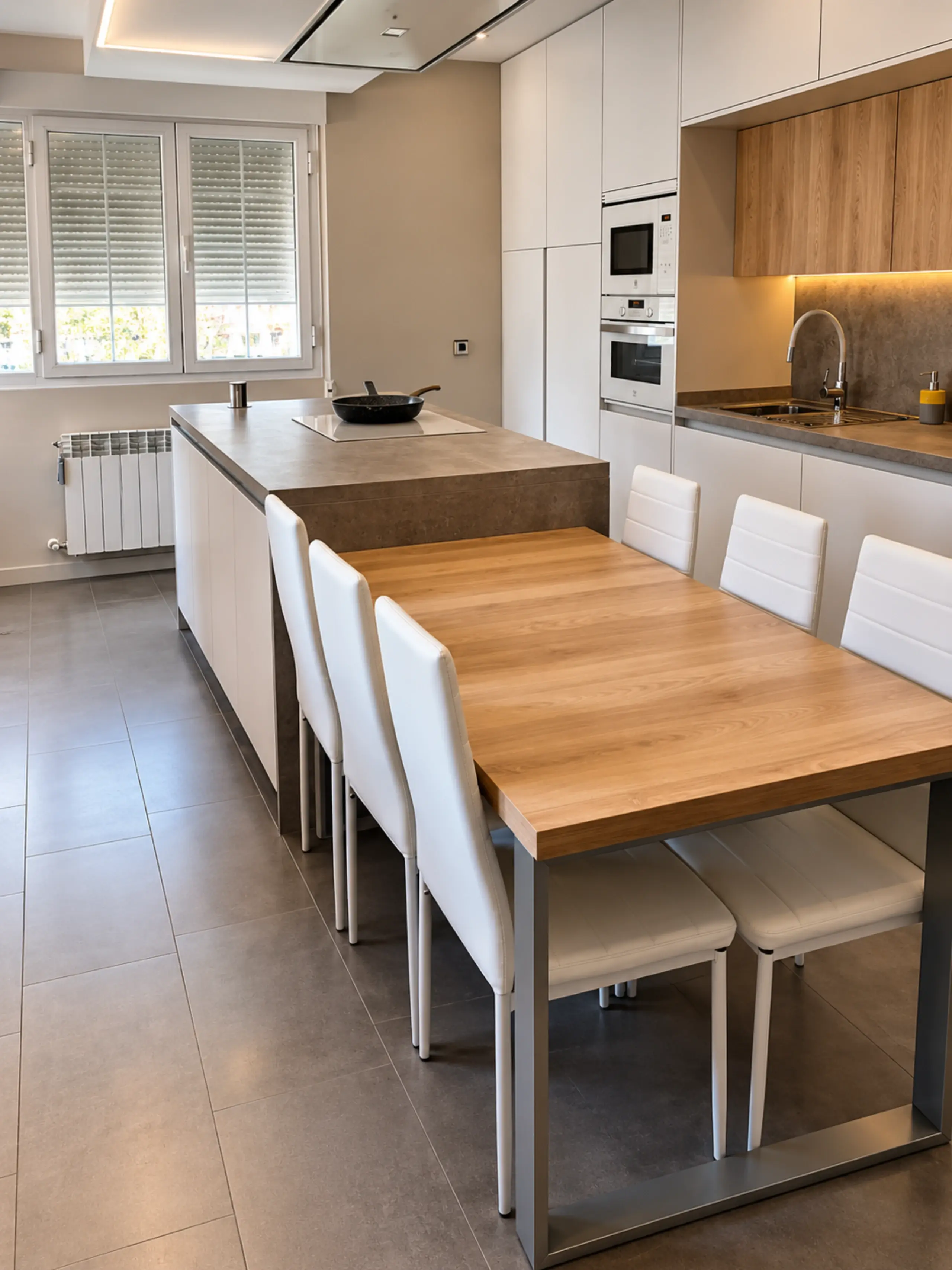 Cocina blanca con muebles altos de madera e isla gris con una mesa de comedor de madera adosada