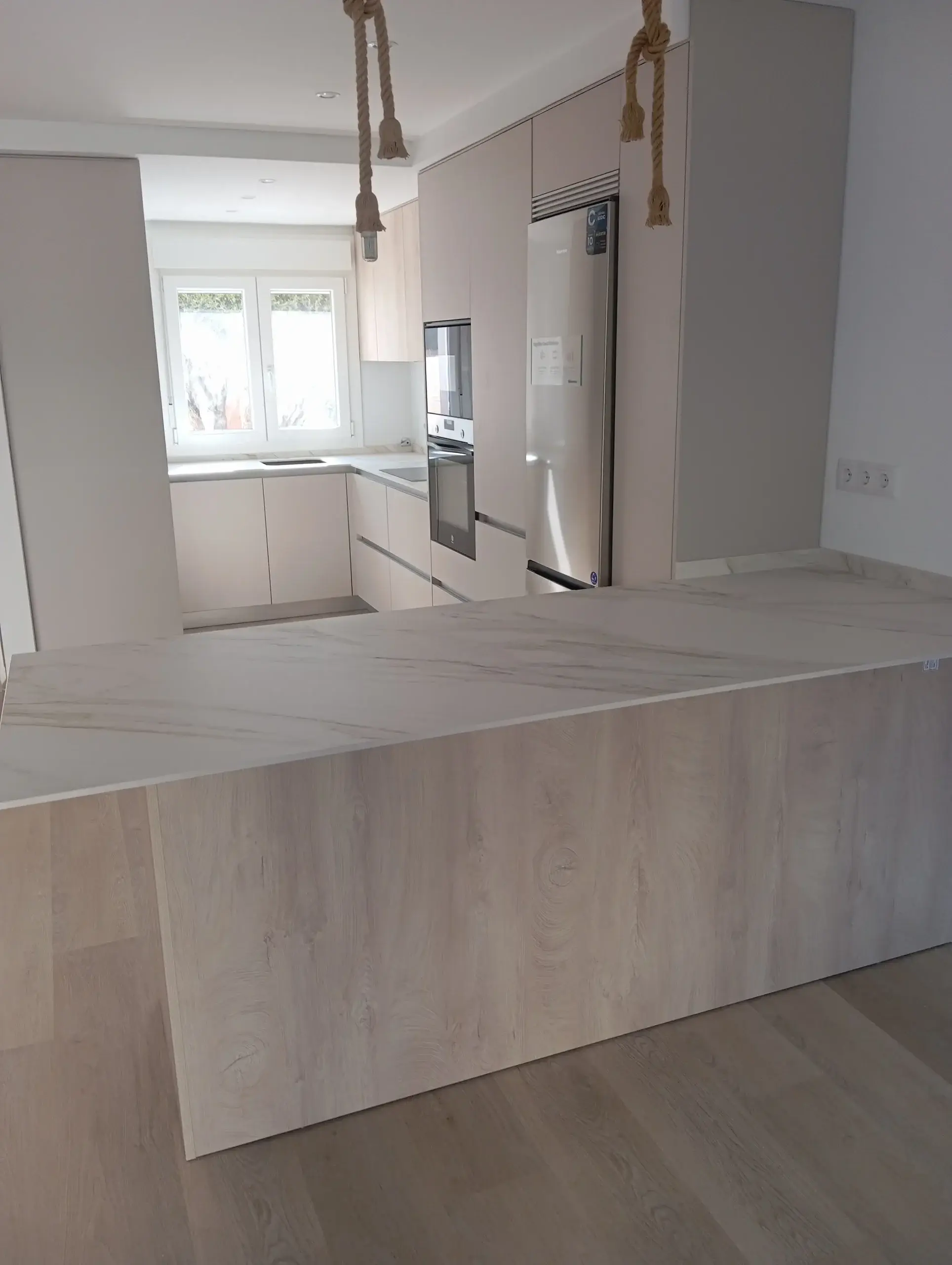 Península de encimera clara que cierra la cocina en G, con columnas y frigorífico al fondo
